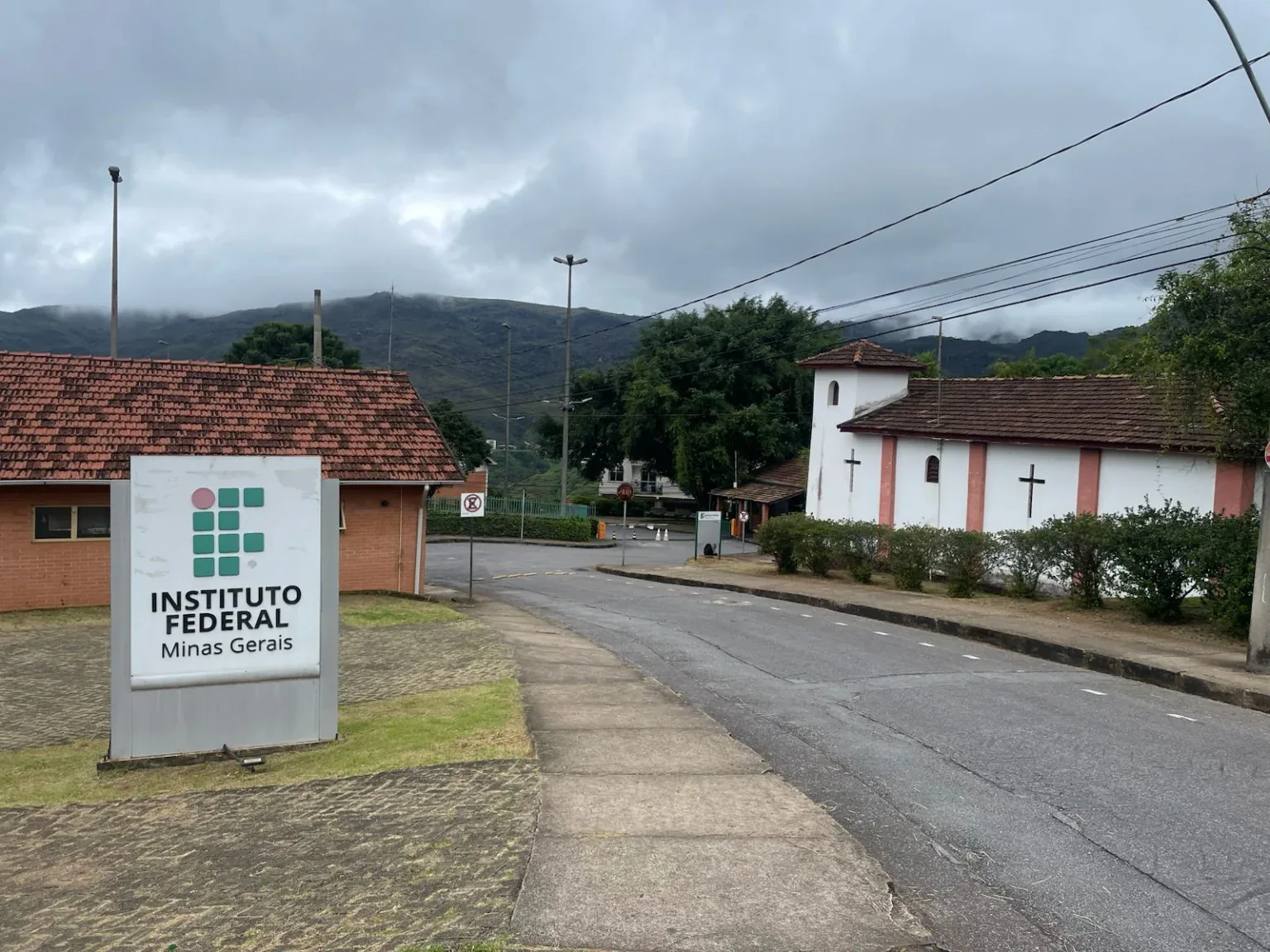Entrada do IFMG campus Ouro Preto, com a placa Instituto Federal Minas Gerais, a capela branca e as serras ao fundo